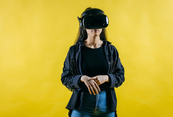 Mod curly dark haired girl dressed in black denim jacket uses the virtual reality glasses on her head and gloves in hands in the studio over yellow background