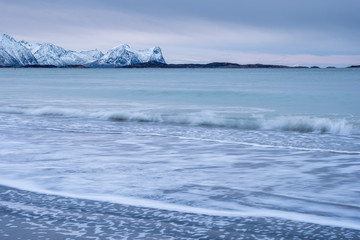 Naklejka na ściany i meble Arctic landscape with long exposure Waves in foreground rolling to coast and snowy mountains in background on a cloudy winter day, Skaland, Senja, Norway