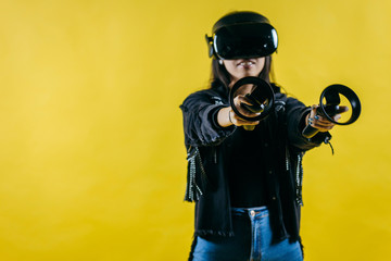 Mod curly dark haired girl dressed in black denim jacket uses the virtual reality glasses on her head and gloves in hands in the studio over yellow background