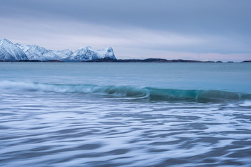 Fototapeta na wymiar Arctic landscape with long exposure Waves in foreground rolling to coast and snowy mountains in background on a cloudy winter day, Skaland, Senja, Norway
