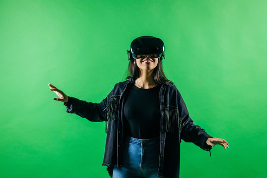 Mod curly dark haired girl dressed in black denim jacket uses the virtual reality glasses on her head and gloves in hands in the studio over green background