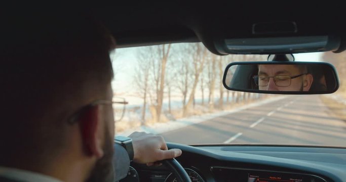 Back view on the Caucasian man in glasses driving a car on the road and looking in the mirror. Rear. View over the shoulder.