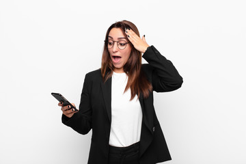 young businesswoman looking happy, astonished and surprised, smiling and realizing amazing and incredible good news and holding a mobile telephone