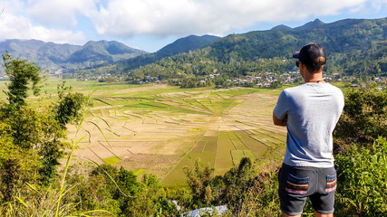 Naklejka premium A man admiring the colorful rice paddies forming a giant spider web in Ruteng, on island of Flores, Indonesia. There are mountains around the paddies. Rural area of Lingko Spider Web Rice Fields