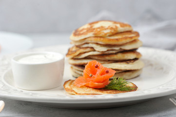 Pancakes with salted salmon.  Fritters served with salmon and sour cream.  Cutlery and a napkin complete the table.  Light background.  Close-up.