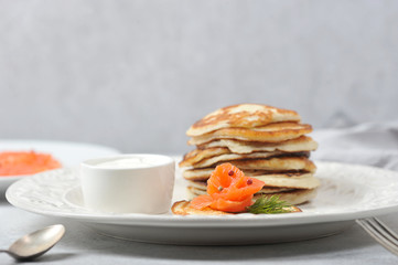 Pancakes with salted salmon.  Fritters served with salmon and sour cream.  Cutlery and a napkin complete the table.  Light background.  Close-up.