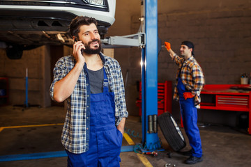 Fototapeta na wymiar smiling mechanic talking on the smartphone with lifted car and coworker on the background