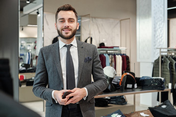 Portrait of positive handsome young businessman with beard holding smartphone in clothing shop