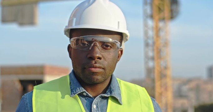 Close up of the African American young handsome man builder in hardhat and goggles looking at the camera and smiling. Outdoor at the building site.
