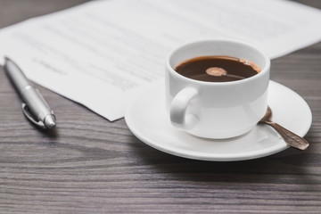 contract signing concept, close-up cup of coffee, silver pen and empty paper sheet of agreement between two counterparties on grey wooden table as background, selective focus