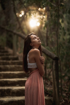 Side view of gentle young in pink dress and white lace bra standing in autumnal park with closed eyes in back lit