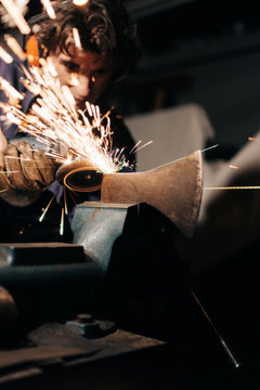 Focused male in gloves and headphones sharpening axe with electric grinder in flying sparkles in workshop