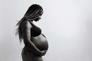 Cheerful young pregnant black female with braids touching belly and looking away with smile while standing against white wall