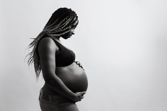 Cheerful young pregnant black female with braids touching belly and looking away with smile while standing against white wall