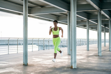 From below African American adult sportswoman in vibrant green activewear focusing and running alone along waterfront among metal columns under roof