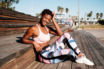 Slim African American young woman in colorful sport clothes and white sneakers looking at camera with curiosity and contemplating while sitting alone on brown wooden stairs on stadium
