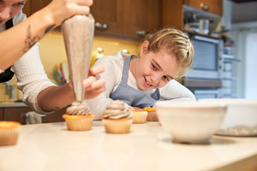 Stock photo of a blonde girl with apron watching as a girl puts cream on top of a cupcake with a pastry sleeve with two children on the side watching