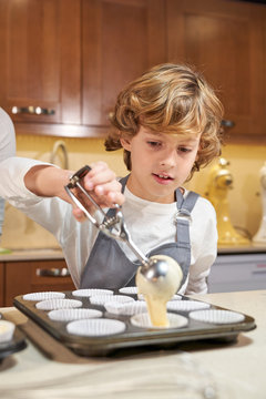 Vertical stock photo of a child with an apron emptying cream on plates ready to make cupcakes