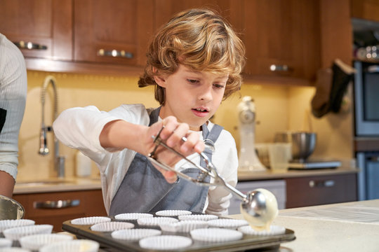 Stock photo of a boy with apron pouring cream on plates prepared to make cupcakes