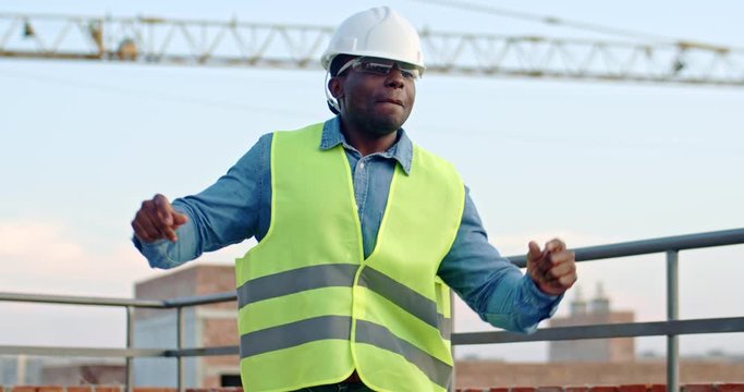 Happy young African American joyful man builder in hardhat dancing and moving funny at the construction site. Outside.