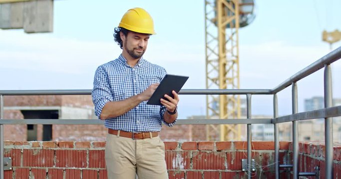 Caucasian young good looking man architect in the hardhat standing at the roof of the building site using tablet device, tapping and scrolling.