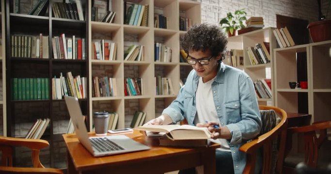 Asian student doing research in library using laptop in books, making a project and preparing for exam. Nerdy guy studying his lessons - education, self-study concept 4k close up