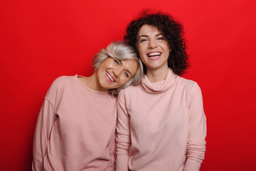 Cute attractive curly woman laughing while her best friend with blonde hair is leaning head on her shoulder laughing dressed in pink against red wall.