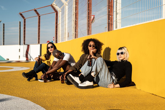 Focused young diverse women in trendy clothes and sunglasses looking at camera while sitting during friendly meeting on stadium