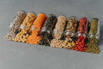 Dried seeds, beans and various grains poured out from glass bottles, spread on grey background....