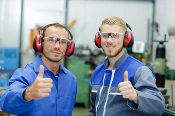 two workers wearing overalls and earmuffs with thumbs up
