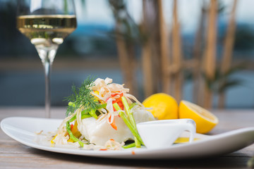 White fish seabass fillet with vegetables and white wine on a wooden table in a restaurant with decor. Tasty and healthy food. A cozy evening in a restaurant during a date. Beautiful table setting.