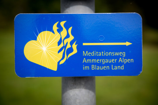 Blue sign in German: Ammergau Alps meditation trail. With a yellow flaming heart