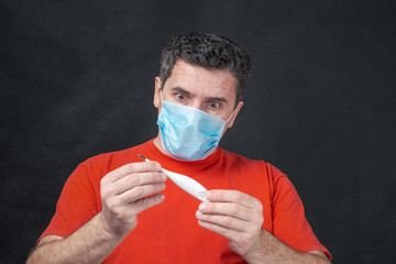 Middle aged caucasian man with medical protective face mask holds thermometer in hands and looking on him with fear. MERS Cov middle East respiratory syndrome coronavirus 2019 nCoV concept