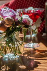 Beautiful little bouquet of undiscovered white pink peonies against the background of a red drink in a can. Buds of flowers stand in a glass little jar on the table in the sunlight, casting a shadow.