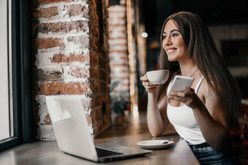 Business woman using phone, working on a laptop and drinking coffee in a cafe.