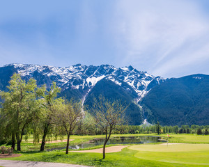 Fototapeta na wymiar Golf course with flag, gorgeous pond and fantastic snow mountain view.