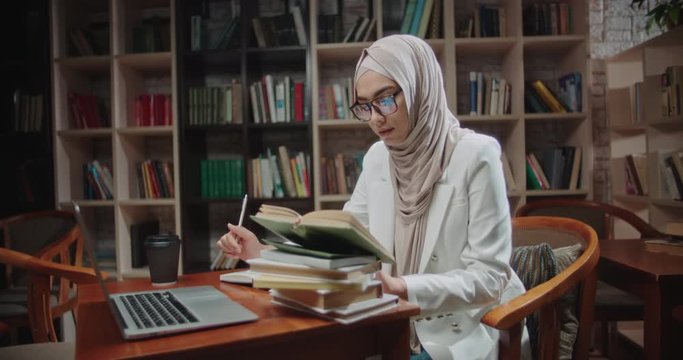 Female muslim student is studying at desk in library, using laptop and books. Girl wearing hijab is preparing for exams - student lifestyle, modern islam concept 4k close up