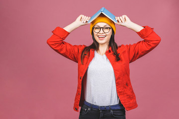 Image of beautiful brunette adult woman wearing eyeglasses smiling and holding book at her head isolated over pink background.