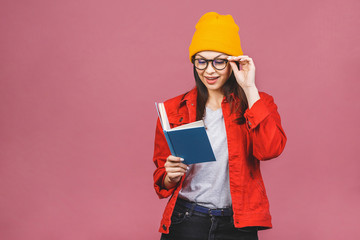 Beautiful young woman wearing casual and eyeglasses standing isolated over pink background, reading a book.