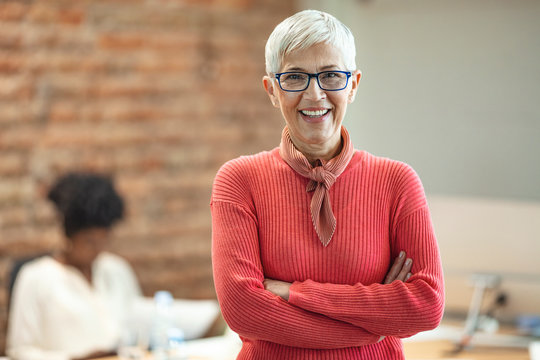 Close-up Portrait Of Mature Business Woman Standing At Office. Pretty Older Business Woman, Successful Confidence With Arms Crossed In Financial Building. Mature Female In Office With Team