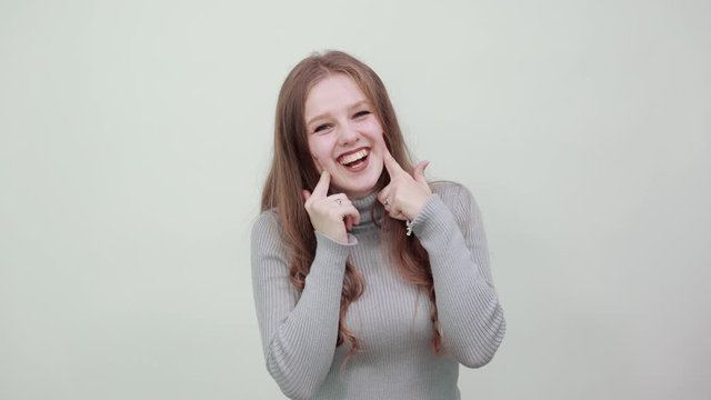 a young beautiful red-haired woman in gray sweater is happy and makes holes in cheeks with index fingers, smiling