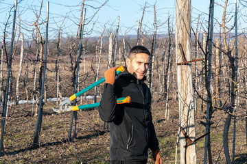 Waist up portrait of the young man in black jacket and orange gloves stands among his fruitgarden, holds shears in hands and looking at the camera, sunny day