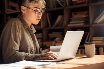 Distance Education. Young woman short hair in glasses sitting at desk studying on laptop browsing...