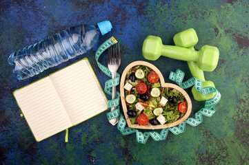 Authentic fresh salad in a wooden heart shaped cup with dumbbells excercise equipment, measuring tape on table. healthy lifestyles, good health