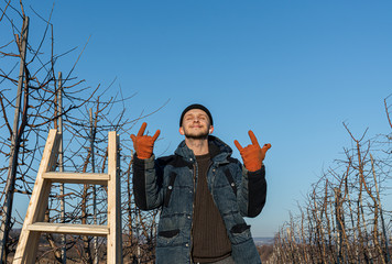 Waist up portrait of the young happy man gardener, dressed in black hat, blue jacket and orange work gloves stands with closed eyes among his garden near the wooden stepladder and shows rock and roll