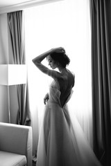 Black and white Portrait of the bride with open dress from the back on the window background. Beautiful neckline on the dress with sexy open back of the bride