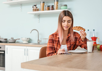 Beautiful young woman with mobile phone having breakfast in kitchen