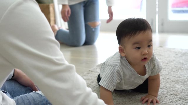 From above of Adorable Asian Infant boy looking at toy in hands of mom and crawling on carpet towards parents while family spending time together at home