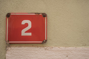 House number two, white letter two or 2 on a red metal plate background, mounted on a yellow wall with four nails.