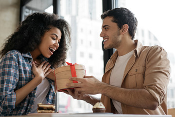 Romantic man giving birthday gift to beautiful emotional woman. Portrait of lovely multiracial couple sitting together in cafe, dating. Valentines day concept